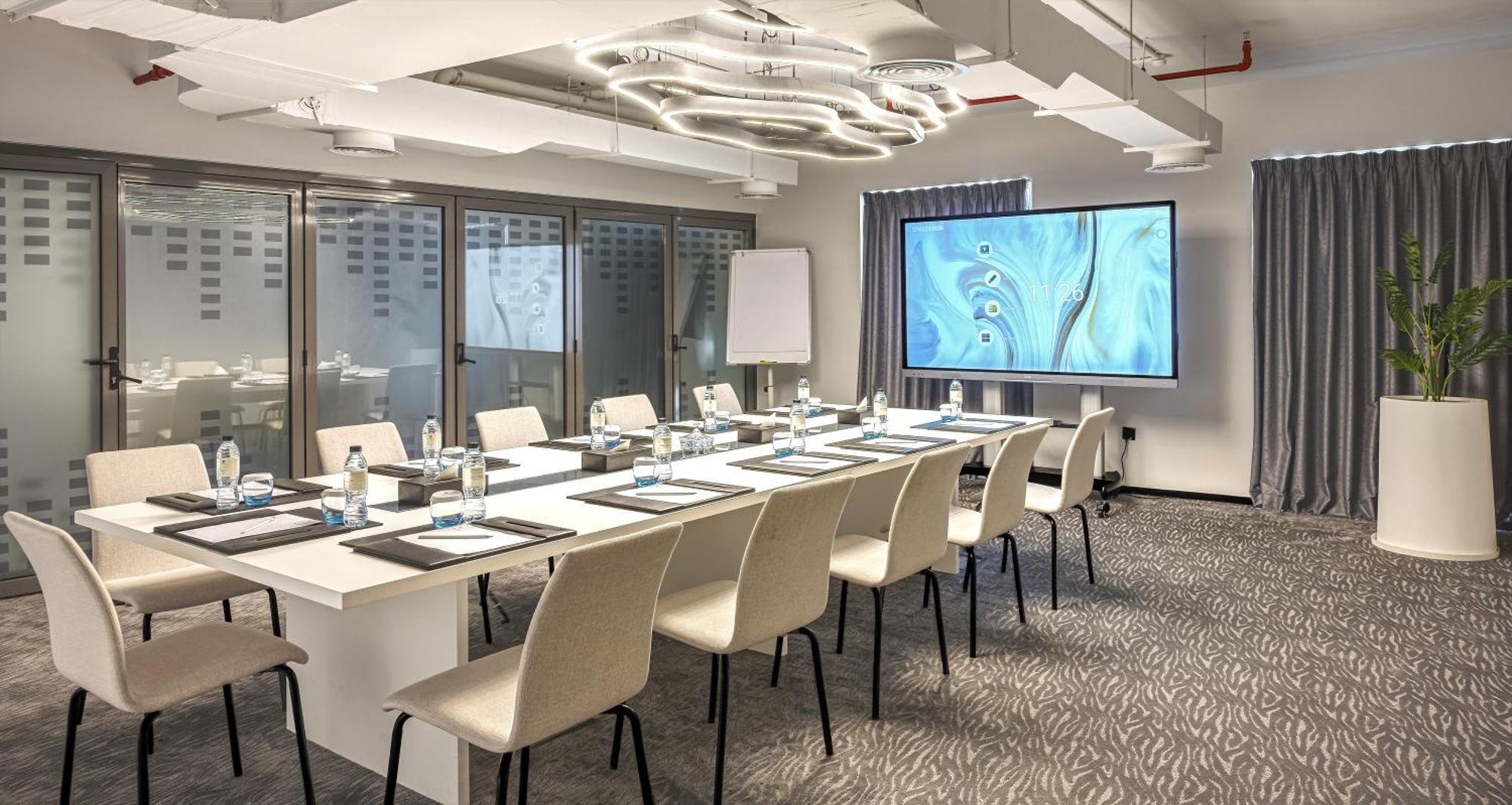 الشقق الفندقية دبيفي  ميلينيوم أتريا بيزنس باي المظهر الخارجي الصورة The photo shows a modern conference room set up for a meeting. There is a long table with several chairs arranged around it, each with a notepad and pen. A large screen displays a graphic or presentation, and there is a whiteboard in the corner. The 