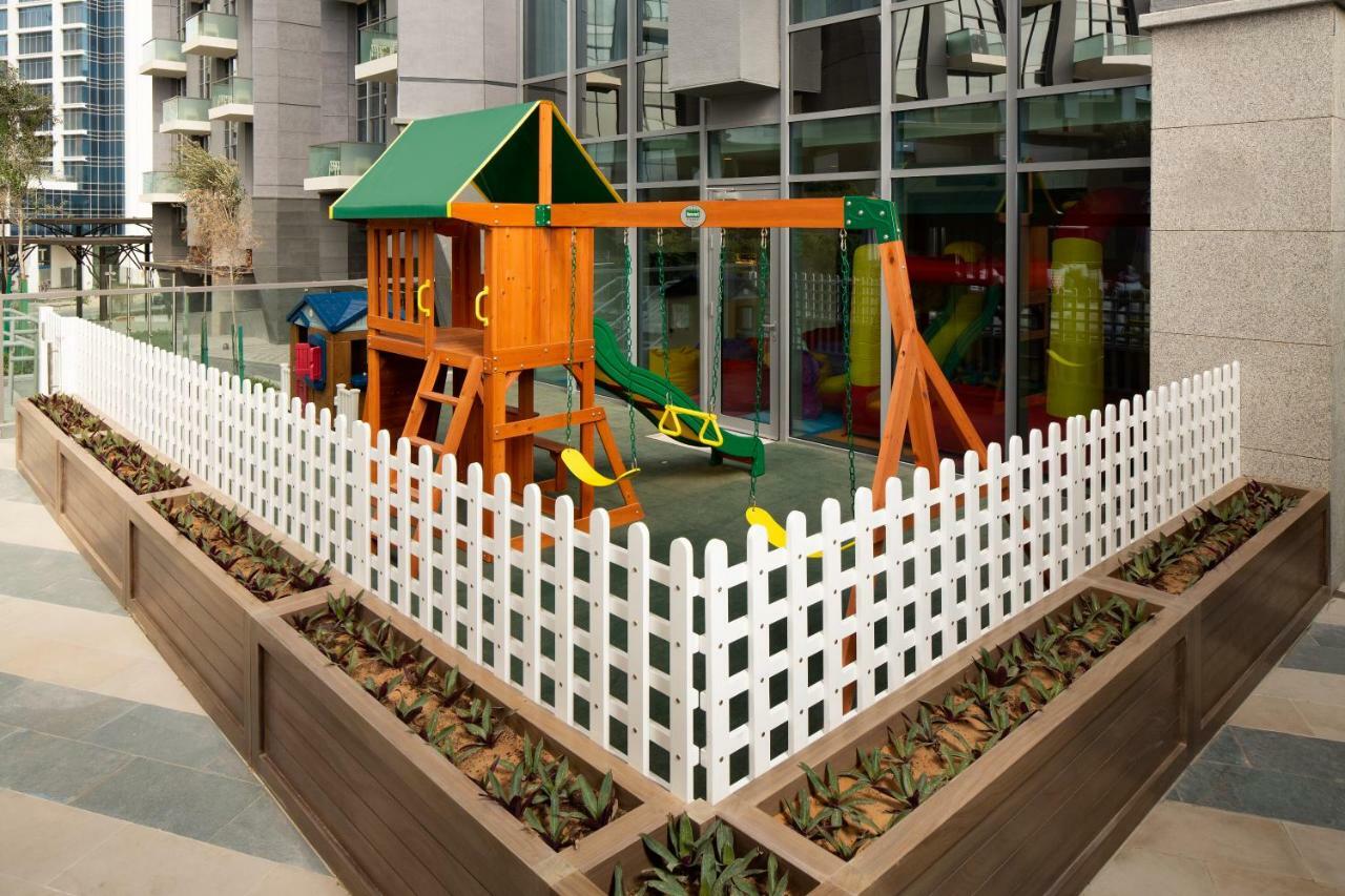 الشقق الفندقية دبيفي  ميلينيوم أتريا بيزنس باي المظهر الخارجي الصورة The image shows a children's playground area, which is enclosed by a white picket fence. The playground features a wooden play structure with a slide, swings, and climbing elements. Surrounding the play area are planters with greenery, and the backdr