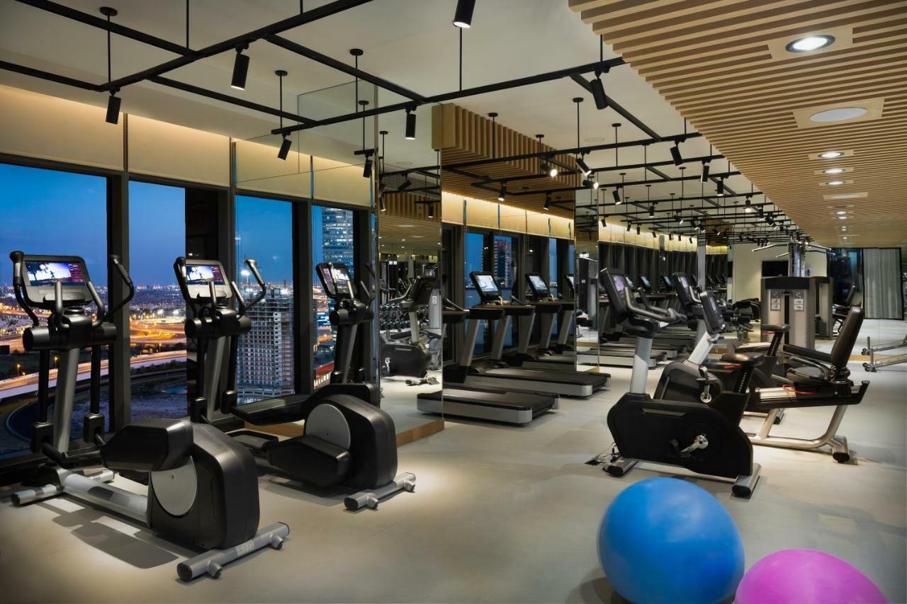 الشقق الفندقية دبيفي  ميلينيوم أتريا بيزنس باي المظهر الخارجي الصورة The photo shows a modern gym with various types of exercise equipment available. There are several treadmills and elliptical machines arranged in rows, with large windows in the background providing a view of the city. The lighting is bright and cont