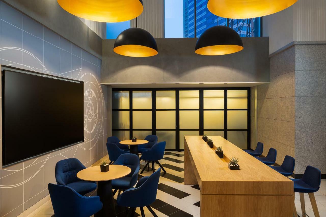 الشقق الفندقية دبيفي  ميلينيوم أتريا بيزنس باي المظهر الخارجي الصورة The photo shows a modern interior space, likely a lounge or meeting area. It features a long wooden table with some small plants on it, accompanied by several blue, circular chairs arranged around it. There is a large flat-screen television mounted o