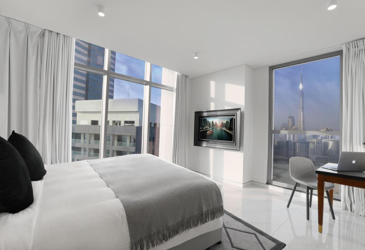 الشقق الفندقية دبيفي  ميلينيوم أتريا بيزنس باي المظهر الخارجي الصورة The photo depicts a modern hotel room that features a large bed with a gray throw blanket. There are large windows allowing natural light to fill the space, showcasing a city skyline view. A flat-screen television is mounted on the wall opposite the 